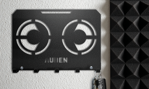 Metallinen seinään kiinnitettävä avainteline, jossa on geometrinen leikkaus ja personoitu nimi, asennettuna teksturoidulle seinälle