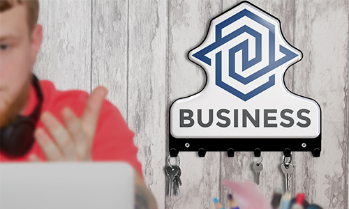 Henkilö punaisessa T-paidassa tietokoneen äärellä, kuulokkeet kaulassa; avainlauta BUSINESS-logolla ja avaimet seinällä