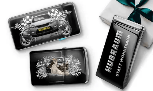 Drei schwarze Geldbörsen mit Auto-Designs und karierten Flaggen, vor einem weißen Hintergrund. Eine zeigt ein Auto mit Rädern, eine andere ein Paar, und eine trägt den Text „Hubraum statt Wohntraum“.