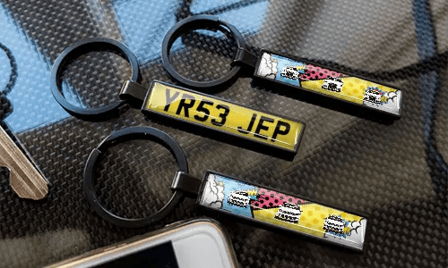 Comic-style license plate keychains displayed on a carbon-textured surface with colorful car designs